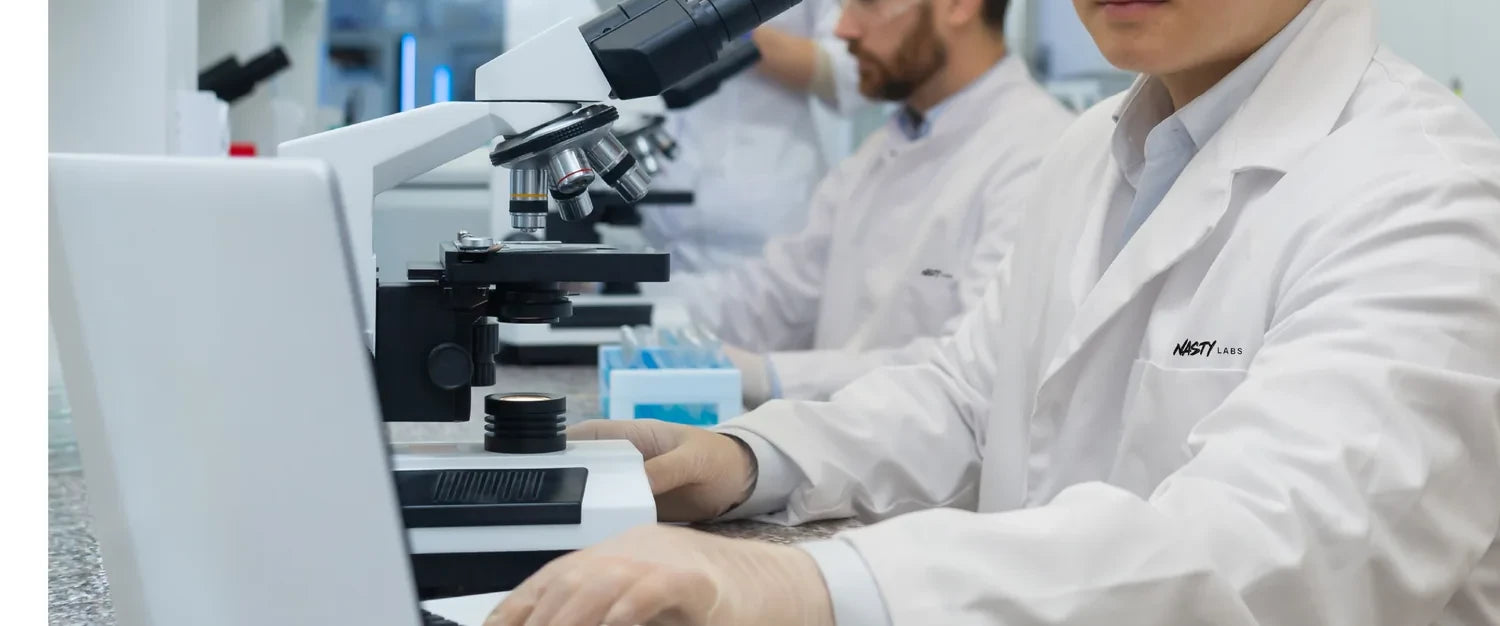 Hands adjusting a microscope on the bench at NASTY Labs, researchers in lab coats working alongside.