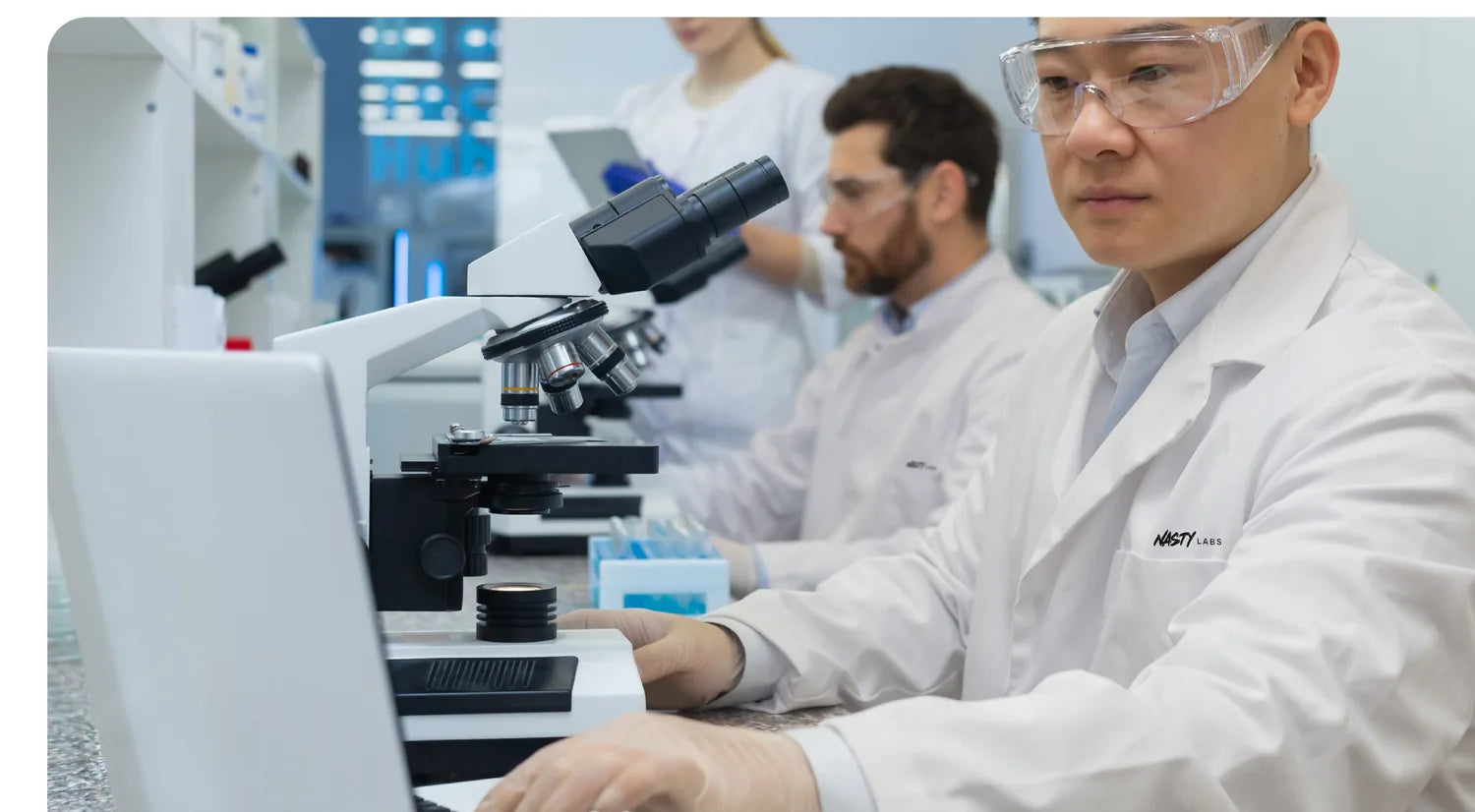 Microscopes, screens and samples on the bench at NASTY Labs, two researchers in lab coats and safety glasses.